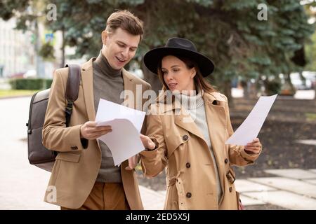 Zwei junge selbstbewusste Kollegen in Smart Casualwear lesen Papiere und Diskutieren sie während der Fahrt entlang der Straße im Park in der Pause Oder nach der Arbeit Stockfoto