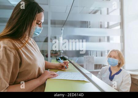 Ärztin oder Krankenschwester, die dem Patienten erklärt, wie er das medizinische Formular an der Rezeption des Krankenhauses ausfüllt. Arzt und Patient tragen Gesichtsschutzmasken und verwenden p Stockfoto