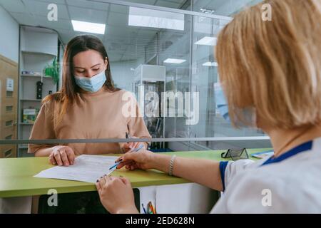 Ärztin oder Krankenschwester, die dem Patienten erklärt, wie er das medizinische Formular an der Rezeption des Krankenhauses ausfüllt. Arzt und Patient tragen Gesichtsschutzmasken und verwenden p Stockfoto