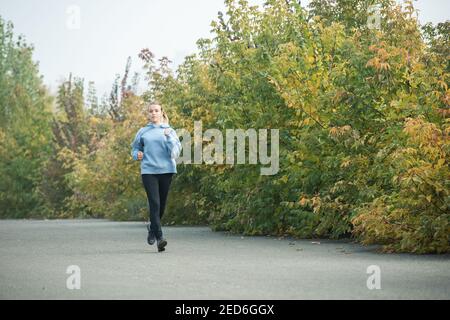 Sportliche und gesunde junge blonde Frau im aktiven Ohr beim Joggen Straße im Park zwischen grünen und gelben Bäumen in der Morgen im Herbst Stockfoto