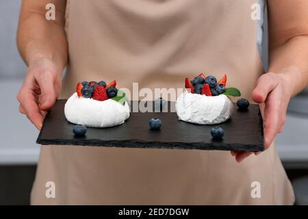 Kuchen Anna Pavlova auf schwarzem Tablett in den Händen des Konditors. Baiser-Kuchen mit frischen Beeren. Stockfoto