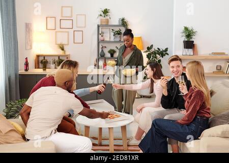 Gruppe von jungen fröhlichen interkulturellen Freunden in Smart Casualwear sitzen am kleinen weißen runden Tisch im Wohnzimmer, mit Pizza und Getränken Stockfoto