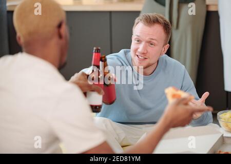 Happy blonde Kerl in hellblau Sweatshirt Blick auf junge Afrikanischer Mann, während beide mit Bierflaschen vorbei klirren Tisch und Snacks Stockfoto