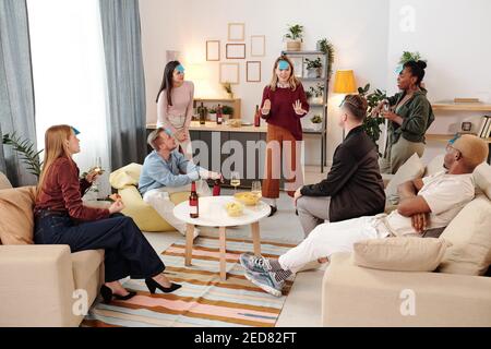 Gruppe von jungen fröhlichen interkulturellen Freunden in smart Casualwear versammelt Um kleinen weißen runden Tisch im Wohnzimmer und spielen Namen Spiel Stockfoto