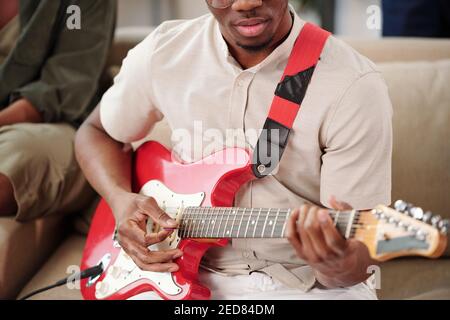 Junge afrikanische Mann in Casualwear spielen E-Gitarre, während sitzen Auf der Couch gegen einen seiner Freunde während der Versammlung bei Zu Hause für Party Stockfoto