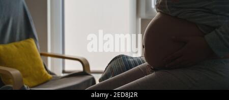 Seitenansicht auf dem Mittelteil der schwangeren kaukasischen Frau, die darauf sitzt Das Bett mit Bett hält Bauch im Schatten gegen die Fenster - Mutterschaft Schwangerschaft conce Stockfoto