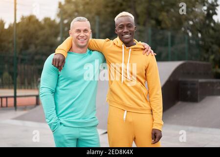Internationale Freundschaft. Zwei glückliche, vielfältige Männer in hellen Sportanzügen stehen im Freien, verbinden sich miteinander und lächeln im Stadtpark mit der Kamera Stockfoto