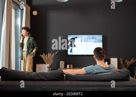 Rückansicht eines jungen, erholsamen Mannes, der vor dem sitzt tv-Gerät und Sport Nachrichten oder Sendung während seiner Frau im Gespräch am Telefon Stockfoto
