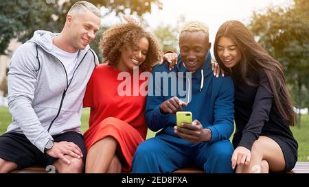 Der junge schwarze Kerl teilt sein Handy, zeigt seinen multiethnischen Freunden in den sozialen Medien einen lustigen Witz und sitzt zusammen auf der Bank im Park Stockfoto