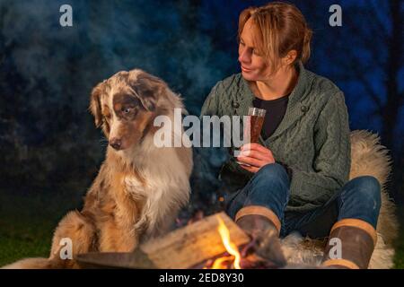 Junge Frau, die am Lagerfeuer sitzt und Wein trinkt. Ihr australischer Schäferhund sitzt neben ihr, sie umarmen sich. Draußen im Park während Stockfoto