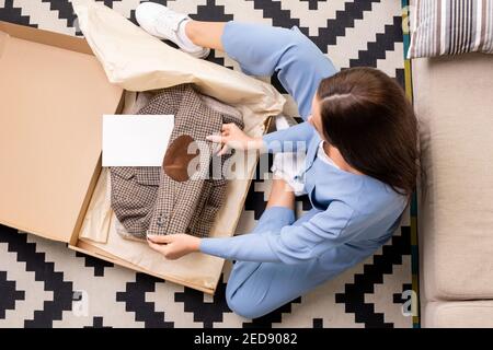 Blick auf junge Frau im blauen Schlafanzug sitzt auf dem Boden im Wohnzimmer, Auspacken und Entfalten karierte Jacke im Online-Shop bestellt Stockfoto