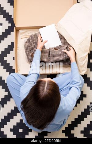Übersicht der jungen Frau in blauen Schlafanzug sitzt auf der Boden und Falten neue karierte Jacke beim Einpacken Papier und Karton Stockfoto