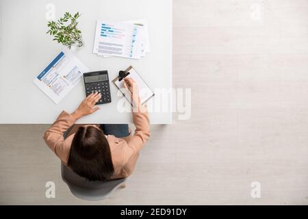 Überblick über junge Buchhalterin mit Taschenrechner am Schreibtisch sitzend, Berechnungen und Aufschreiben von Informationen auf Papier Stockfoto