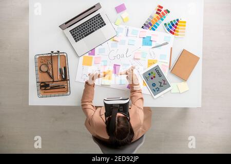 Draufsicht der kreativen Frau mit vr-Headset Vorbereitung der Präsentation, während Sie am Schreibtisch mit mobilen Gadgets, Paletten und Bürobedarf sitzen Stockfoto