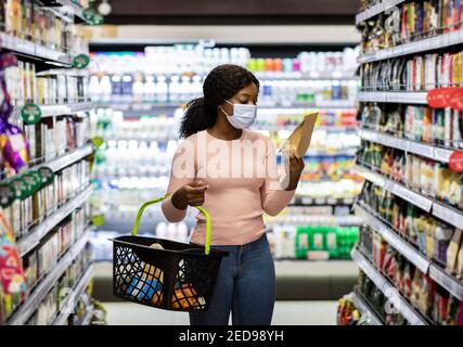 Junge schwarze Frau in Gesichtsmaske Einkaufen für Lebensmittel bei Riesiger Supermarkt während der covid-19 Sperre Stockfoto