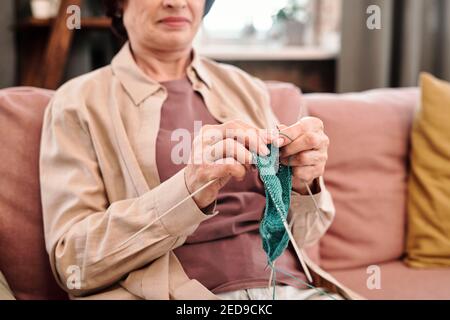 Hände von glücklich reifen Frau in Casualwear sitzen auf weich Bequeme Couch im Wohnzimmer und Strickschal oder Socken für Ihre Enkel Stockfoto