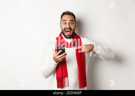 Aufgeregt Mann erzählt über Angebot auf Handy, zeigt auf Smartphone und schaut erstaunt, stehen vor weißem Hintergrund Stockfoto