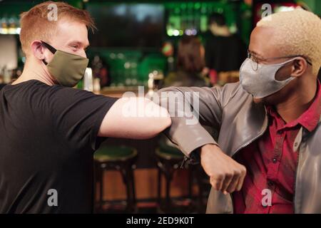 Zwei junge interkulturelle Männer in Casualwear und Schutzmasken suchen Beim Ellenbogenstoß gegen den Stangenzähler ineinander Modernes Pub Stockfoto