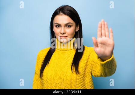 Unzufriedene kaukasische junge Brünette Frau in gelben Casual Pullover, zeigt Stop-Geste mit Palm suchen Kamera, steht auf isolierten blauen Hintergrund. Stockfoto