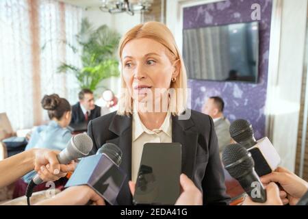 Reife blonde weibliche Delegierte in formalwear stehen vor Journalisten mit Mikrofonen und beantworten ihre Fragen im Interview Stockfoto