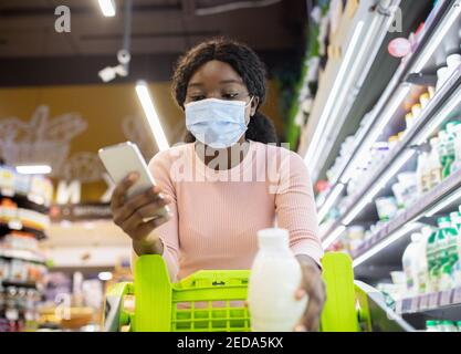 Junge schwarze Frau in Gesichtsmaske Blick in ihr Lebensmittelgeschäft Smartphone-App beim Einkaufen im Milchbereich des Supermarkts Stockfoto