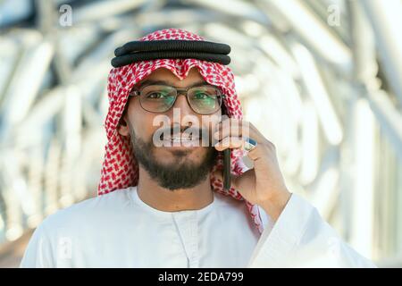 Lächelnder junger arabischer Delegierter oder Geschäftsmann in nationaler Kleidung im Gespräch Auf dem Mobiltelefon vor der Kamera im modernen Business Zentrieren Stockfoto