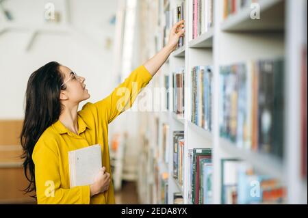 Kaukasische Studentin steht in der Universitätsbibliothek, auf der Suche nach einem Buch. Schöne junge Frau in Brillen auf der Suche nach Informationen für ein Projekt, Lernen für bestandene Prüfungen, Bildungskonzept Stockfoto