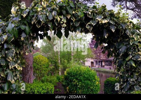 BOURTON-ON-THE-WATER, ENGLAND - APRIL 28th 2019 : Blick auf den Fluss Windrush und einige Geschäfte in Cotswold Stein durch einen Efeubogen Stockfoto