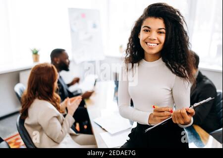 Porträt einer eleganten Managerin bei einem Geschäftstreffen. Erfolgreiche junge Geschäftsfrau schaut direkt auf die Kamera, lächelt. Schöne Dame in formeller Kleidung bei Briefing im modernen Büro Stockfoto