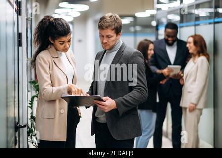 Multiethnische Kollegen stehen im Büro. Schöne afroamerikanische weibliche Angestellte konsultiert kaukasischen männlichen Kollegen auf Arbeitsprozess. Fokussierte junge Erwachsene Frau und Mann diskutieren das Projekt Stockfoto