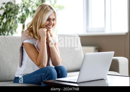 Eine zufriedene blonde Dame mittleren Alters sitzt zu Hause im Wohnzimmer, schaut auf den Laptop-Bildschirm und lächelt glücklich, erhielt unerwartete Nachrichten oder eine gute Nachricht Stockfoto