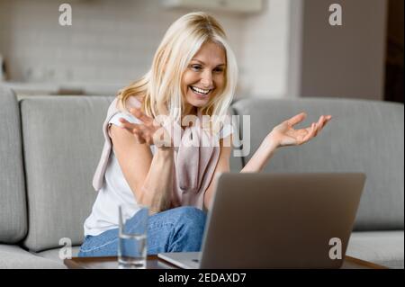 Glücklich lächelnde kaukasische ältere Frau mit Brille sitzt auf der Couch im Wohnzimmer, mit Laptop, während der Arbeit aus der Ferne, kommuniziert mit Geschäftskollegen Stockfoto