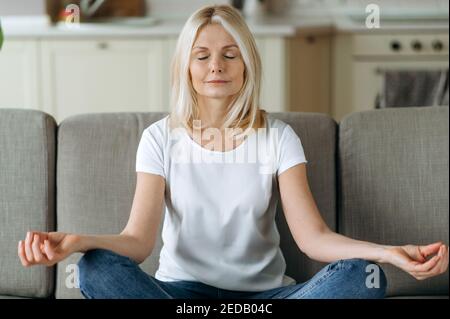Ruhige attraktive Frau mittleren Alters sitzt auf dem Sofa zu Hause und chillen. Ältere Frau ist Praxis Yoga und Meditation in Lotusposition mit geschlossenen Augen, Stress zu lindern, Ruhe Konzept Stockfoto