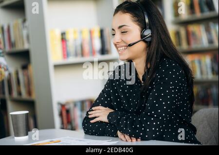 Lächelnde junge kaukasische Frau, Manager, Tutor oder Callcenter-Betreiber, sitzt an der Rezeption, sprechen per Videoanruf mit Kollegen oder Studenten. Zufriedene Frau mit Headset sprechen online per Videoanruf Stockfoto