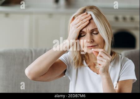 Verärgert ungesunde kaukasische reife Frau zu Hause auf dem Sofa sitzen Trinken Schmerzlinderung Pille für Kopfschmerzen mit geschlossenen Augen Stockfoto