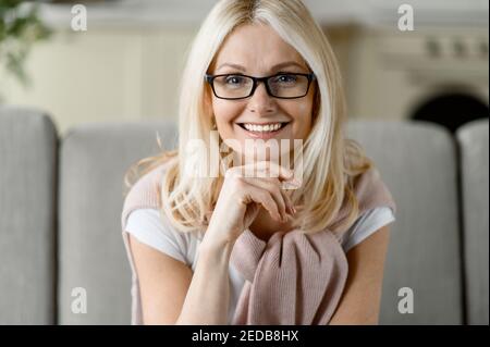 Nahaufnahme Porträt einer kaukasischen blonden Frau mittleren Alters, die eine Brille trägt, auf der Couch sitzt und mit freundlichem Lächeln direkt auf die Kamera blickt Stockfoto