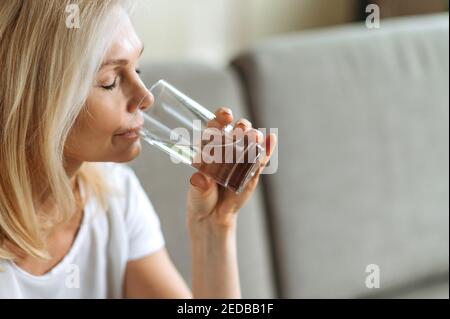 Folgen Sie einem gesunden Lebensstil. Nahaufnahme einer reifen kaukasischen attraktiven Frau, sitzt auf dem Sofa zu Hause, genießt ein Glas sauberes Wasser mit geschlossenen Augen trinken, die tägliche Norm Stockfoto