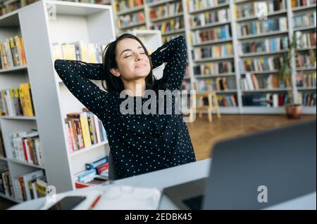 Glückliche weibliche Angestellte, die im modernen Büro sitzt, mit den Händen hinter dem Kopf. Stilvolle Managerin, die eine Pause von der Arbeit macht und sich nach einem Brainstorming ausruhen kann. Selbstbewusste junge Dame am Arbeitsplatz entspannen Stockfoto