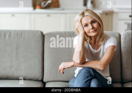Attraktive ruhige kaukasische Frau mittleren Alters sitzt auf dem Sofa im Wohnzimmer in lässiger Kleidung gekleidet, schaut in die Kamera und lächelt, Rest von Hausaufgaben, Wochenende Stockfoto