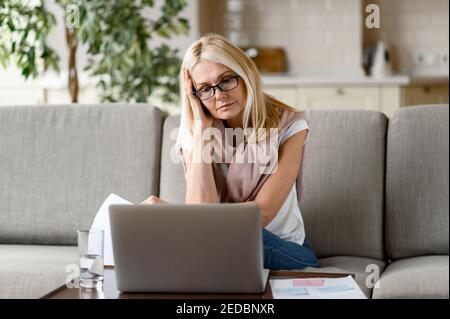 Fokussierte müde Senior blonde arbeitet zu Hause aus der Ferne, sitzt auf der Couch im Wohnzimmer, analysiert monatliche Berichte, verwendet einen Laptop. Arbeiten Sie von zu Hause aus Stockfoto