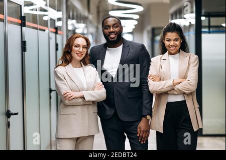 Multirassische einflussreiche Geschäftsleute in formeller Kleidung schauen lächelnd auf die Kamera. Glückliche weibliche und männliche Kollegen stehen im modernen Büro, zeigen Vertrauen und Erfolg, Teamwork-Konzept Stockfoto