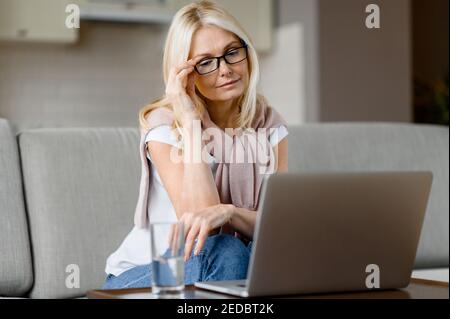 Ältere ruhige kaukasische Frau sitzt auf der Couch zu Hause im Wohnzimmer, mit Laptop, Surfen im Internet, Online-Shopping, Remote arbeiten Stockfoto