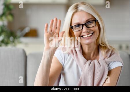 Webcam-Porträt einer glücklichen reifen kaukasischen blonden Frau in Brillen, auf der Couch sitzend, direkt auf die Kamera mit freundlichem Lächeln, winkende Hand, Grußgeste Stockfoto