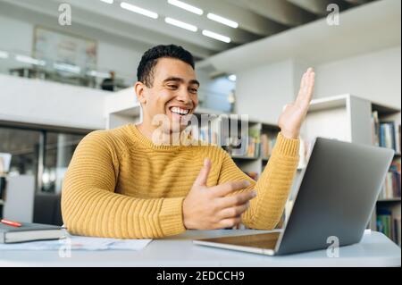 Gutaussehender männlicher Angestellter während einer Videokonferenz mit Kollegen. Hispanic Freiberufler oder Student sitzt am Schreibtisch arbeiten oder lernen mit Laptop und lächeln Stockfoto