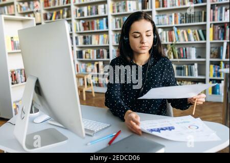 Fokussierte Geschäftsfrau macht einen Papierkram, sitzt am Schreibtisch im Büro. Kaukasische Angestellte, die ein Headset trägt, als Callcenter-Telefonanbieters oder als Tutorin im Unternehmen arbeitet Stockfoto