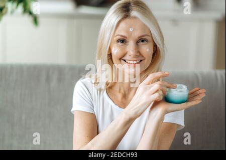 Lächelnde blonde ältere Frau tragen Anti-Aging-Creme auf ihr Gesicht auf. Glückliche reife Frau mit kosmetischen Creme Falten im Gesicht zu verstecken, spendet Feuchtigkeit und pflegt die Haut Stockfoto