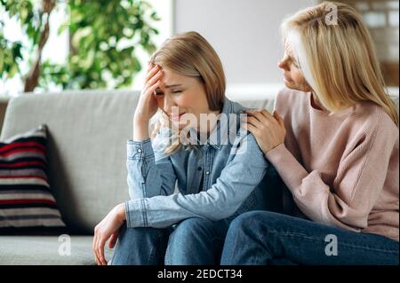 Besorgt Mutter mittleren Alters umarmt und Konsolen junge Erwachsene Tochter, sitzt auf dem Sofa, ältere Mutter beruhigen weinende erwachsenen Kind. Unterstützung eines geliebten Menschen, eines Kindes. Stockfoto