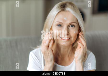 Pflege der alternden Haut. Attraktive Kaukasierin mittleren Alters kümmert sich um ihr Gesicht, wendet Anti-Aging-Faltencreme an, schaut auf die Kamera und lächelt. Nahaufnahme Stockfoto