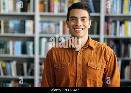 Portrait des erfolgreichen hispanischen Geschäftsmanns schaut direkt auf die Kamera, lächelt. Glückliche männliche Freiberufler oder Student im modernen Büro oder Universitätsbibliothek stehen Stockfoto