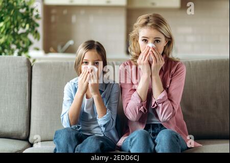 Traurige kaukasische Mutter und Tochter mit Erkältung sitzen zu Hause auf dem Sofa mit Servietten, niesen, husten, laufende Nase. Erkältung, Grippe, Krankheit Stockfoto